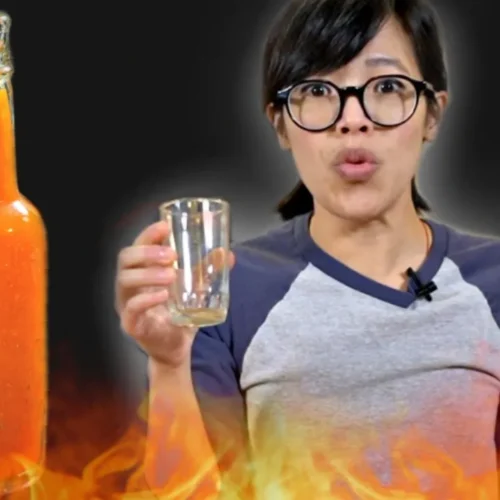 Emmy holding empty glass with flames surrounding her. Not sauce bottle fills frame to her right.