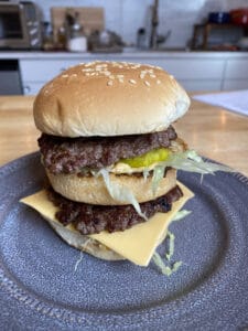 Homemade Big Mac on a plate. From top to bottom: sesame bun, beef patty, shredded lettuce, sauce, middle bun, second patty, cheese bottom bun.